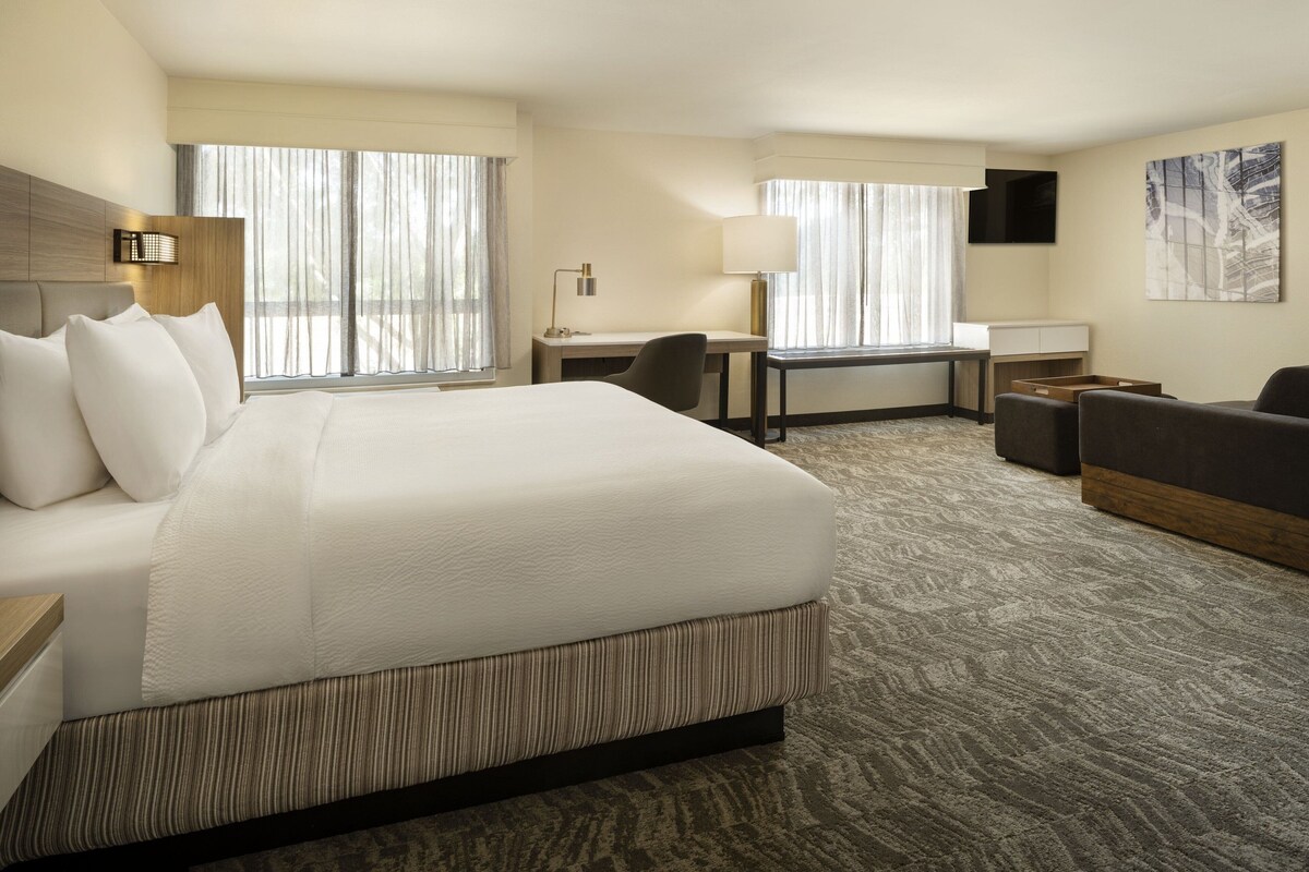 A hotel room with a large bed, a desk, a chair, a sofa, and a TV on the wall.