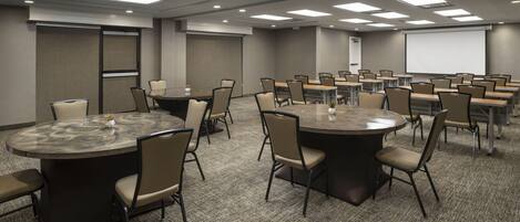 A conference room with multiple round tables and chairs, a projector screen, and a carpeted floor.