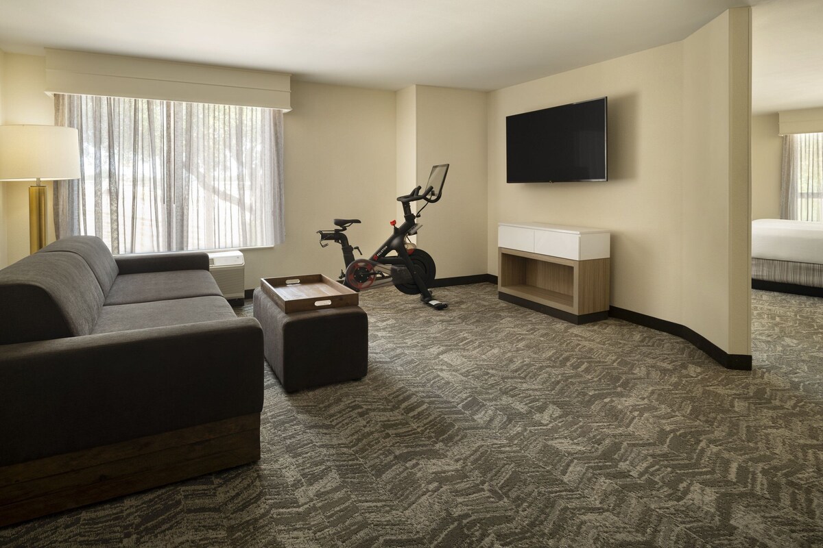 A hotel room with a sofa, a small table, an exercise bike, a TV, and a bed.