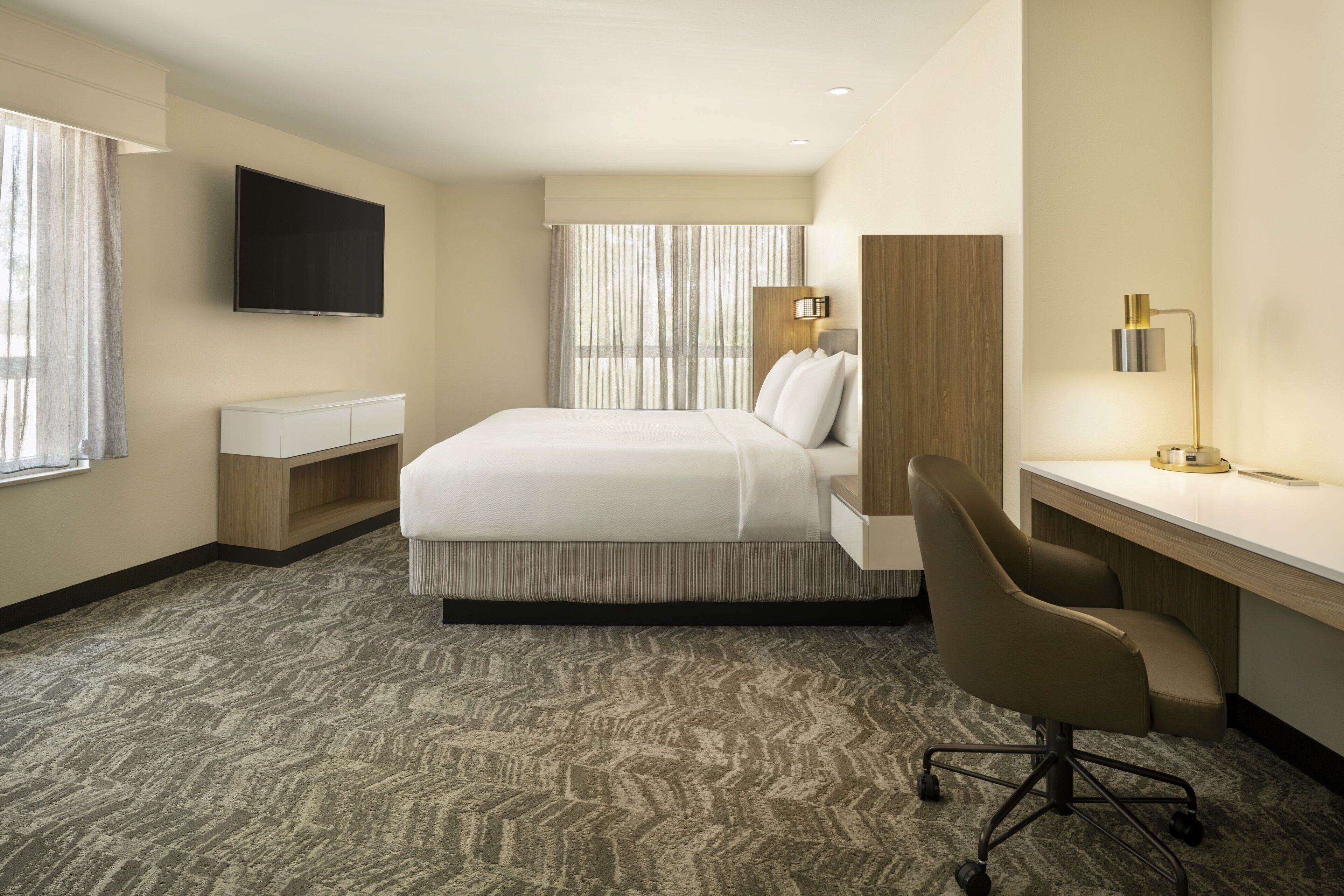 Habitación de hotel con cama, escritorio, silla, televisor y ventana con cortinas.