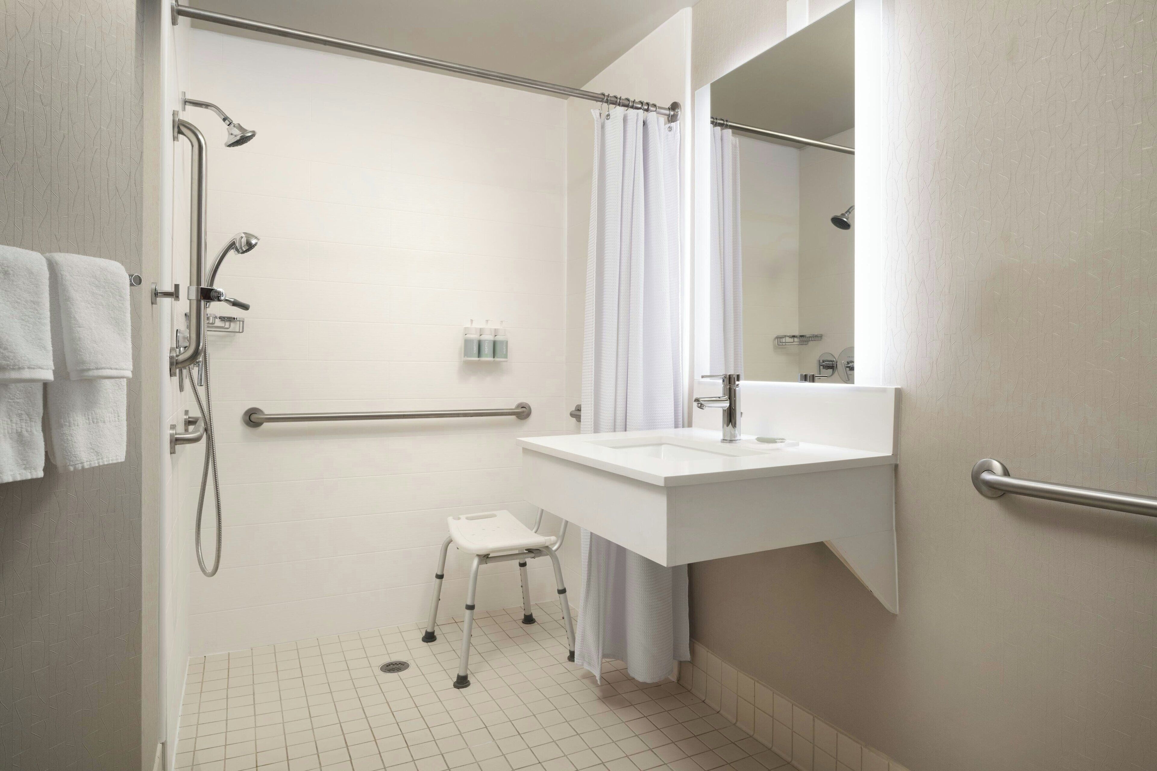 A bathroom with a shower, grab bar, white sink, and a small stool.