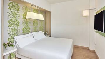 A neatly made bed in a hotel room with a green floral wallpaper, a bedside table with a vase of flowers, and a TV mounted on the wall.