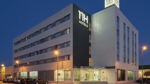 A modern hotel building with a prominent 'NH Hotels' sign, a ground-level entrance, and a parking area with several cars. - NH Campo Cartagena (Cartagena)