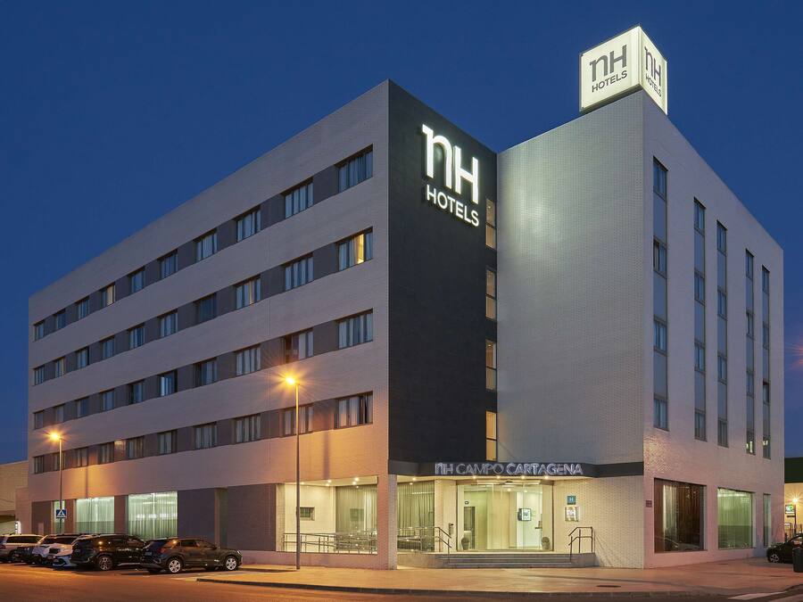 A modern hotel building with a prominent 'NH Hotels' sign, a ground-level entrance, and a parking area with several cars.
