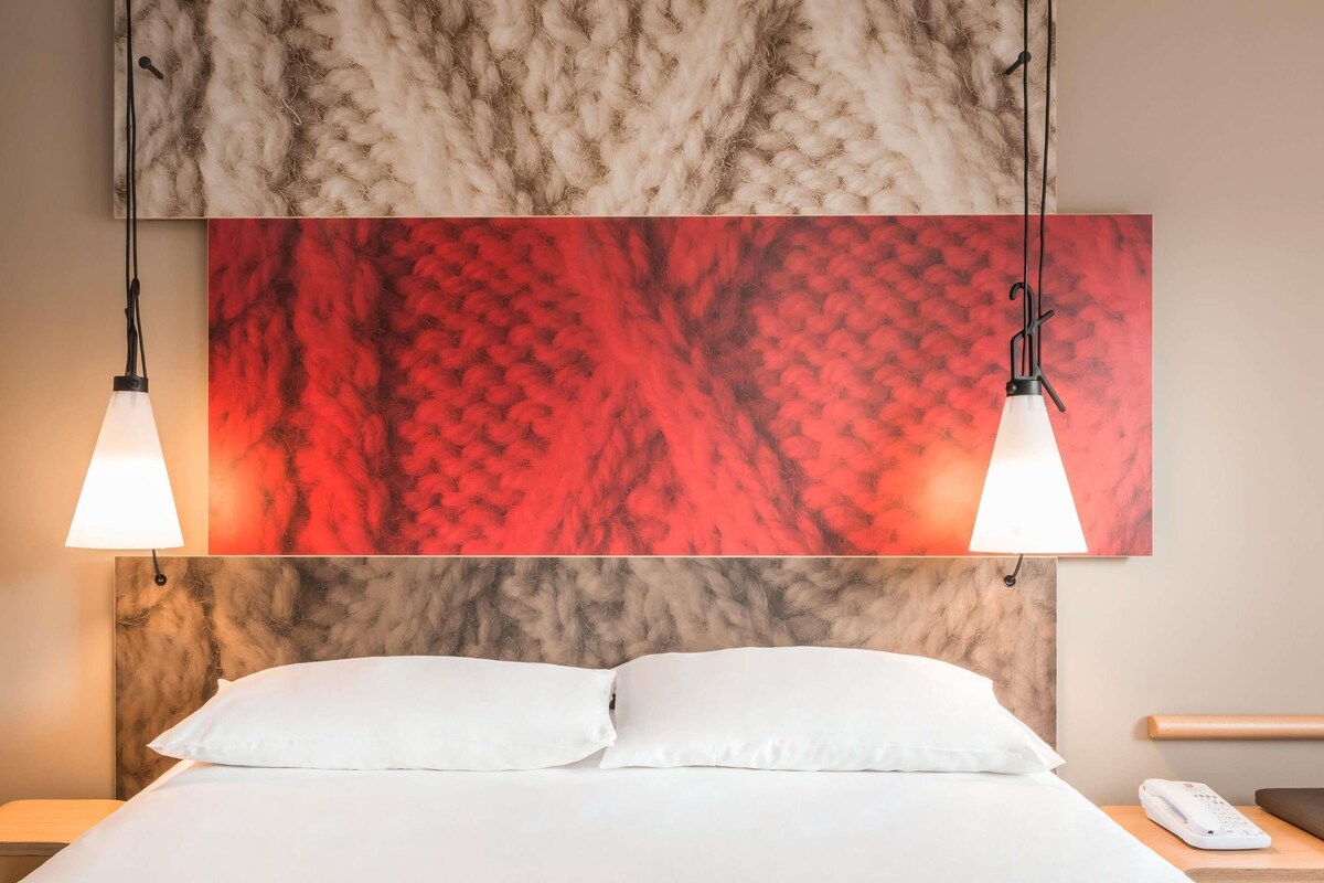 A hotel room with a bed, two pendant lights, a red and beige textured wall art, a white pillow, a bedside table with a phone, and a wooden nightstand.