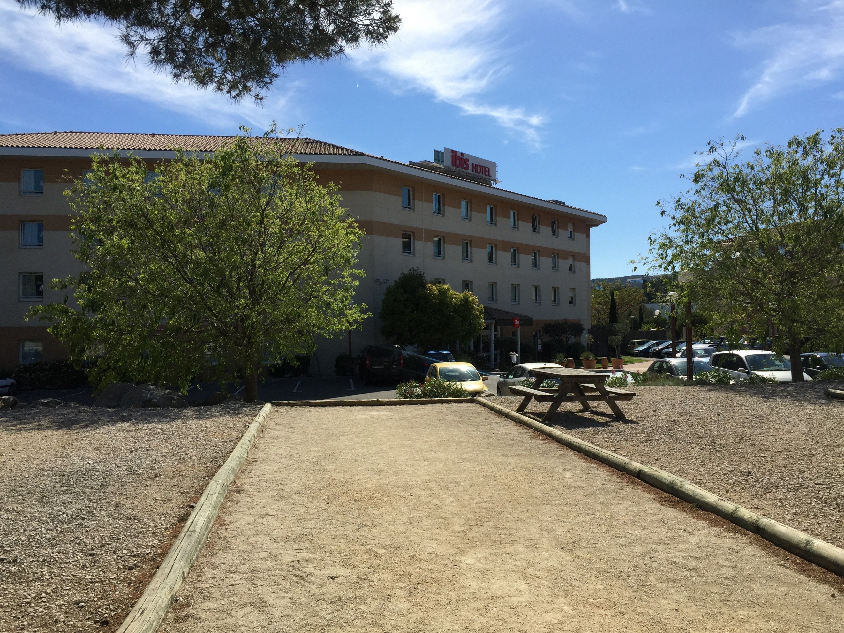 Il s’agit d’un hôtel avec un toit recouvert de tuiles, de nombreuses fenêtres et une enseigne indiquant qu’il s’agit d’un hôtel Ibis. On y trouve un parking avec plusieurs voitures et une table de pique-nique.