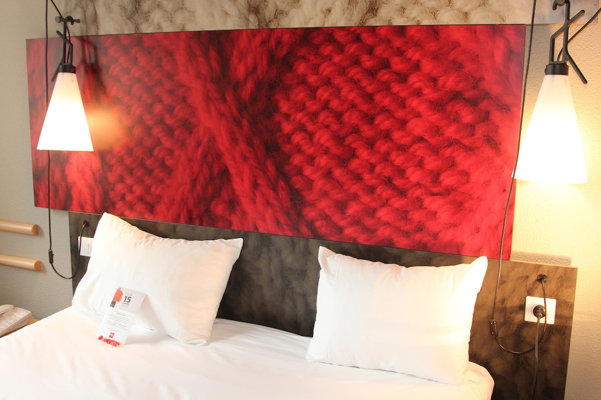 A hotel room with a large red textured wall, two white pillows, a white bedspread, and two pendant lights.