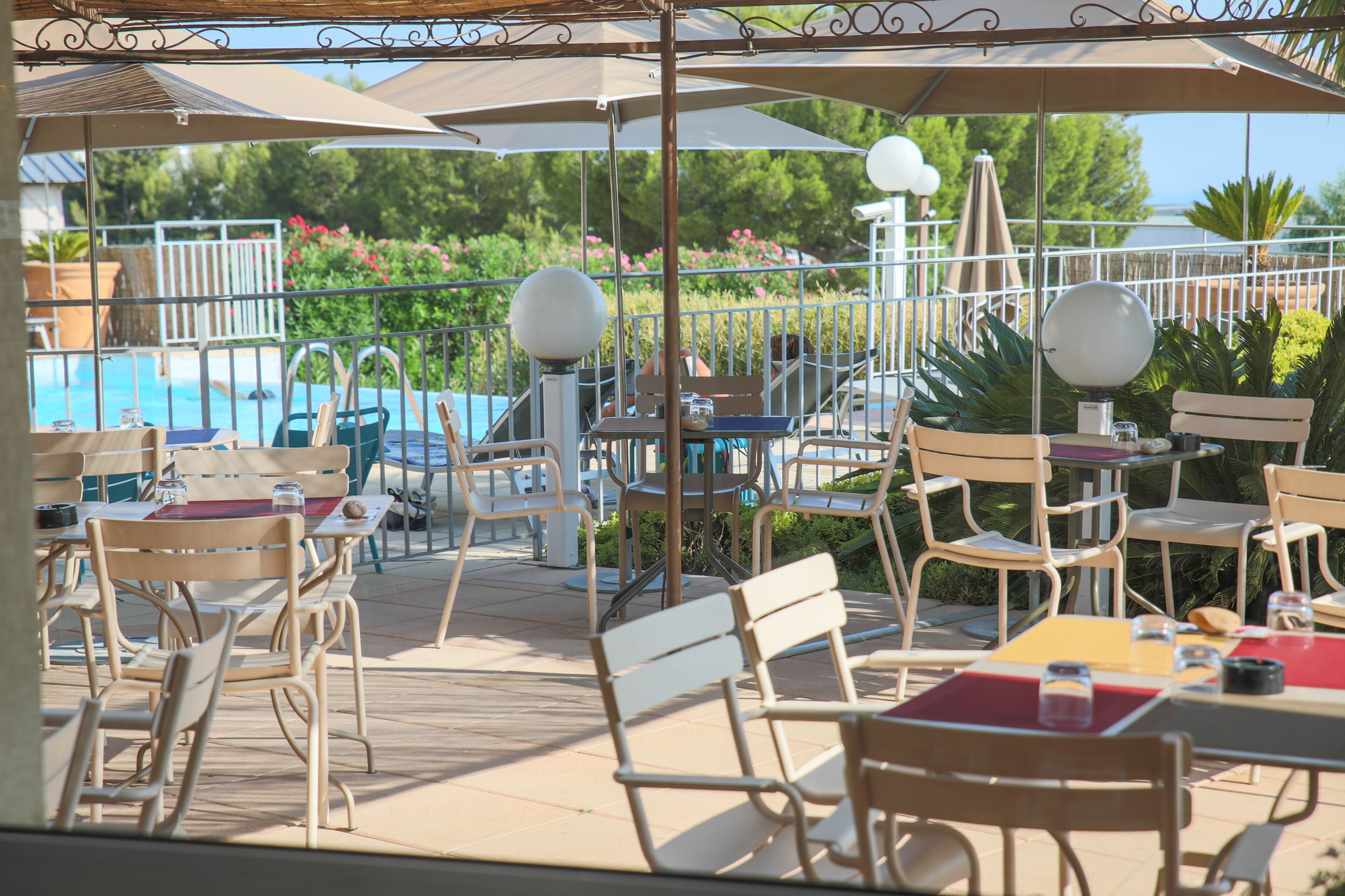 Un espace extérieur aménagé en terrasse, avec des tables et des chaises, des parasols, et une piscine en arrière-plan.