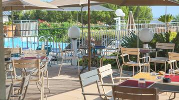 Un espace extérieur aménagé en terrasse, avec des tables et des chaises, des parasols, et une piscine en arrière-plan.