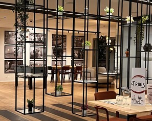 A modern restaurant with a central dining area, surrounded by black metal partitions with plants. Tables are set with wine glasses and menus. - ibis Fribourg (Granges-Paccot)