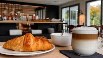 A croissant on a plate, a bowl with a spoon, and a glass of coffee on a table in a modern cafe setting.