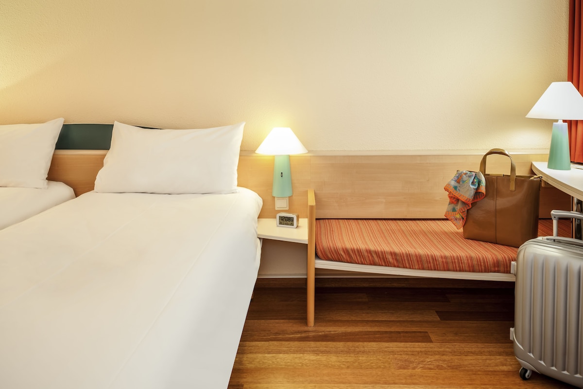 Een hotelkamer met twee bedden, een nachtkastje met een lamp, een koffer en een handtas op de vloer.