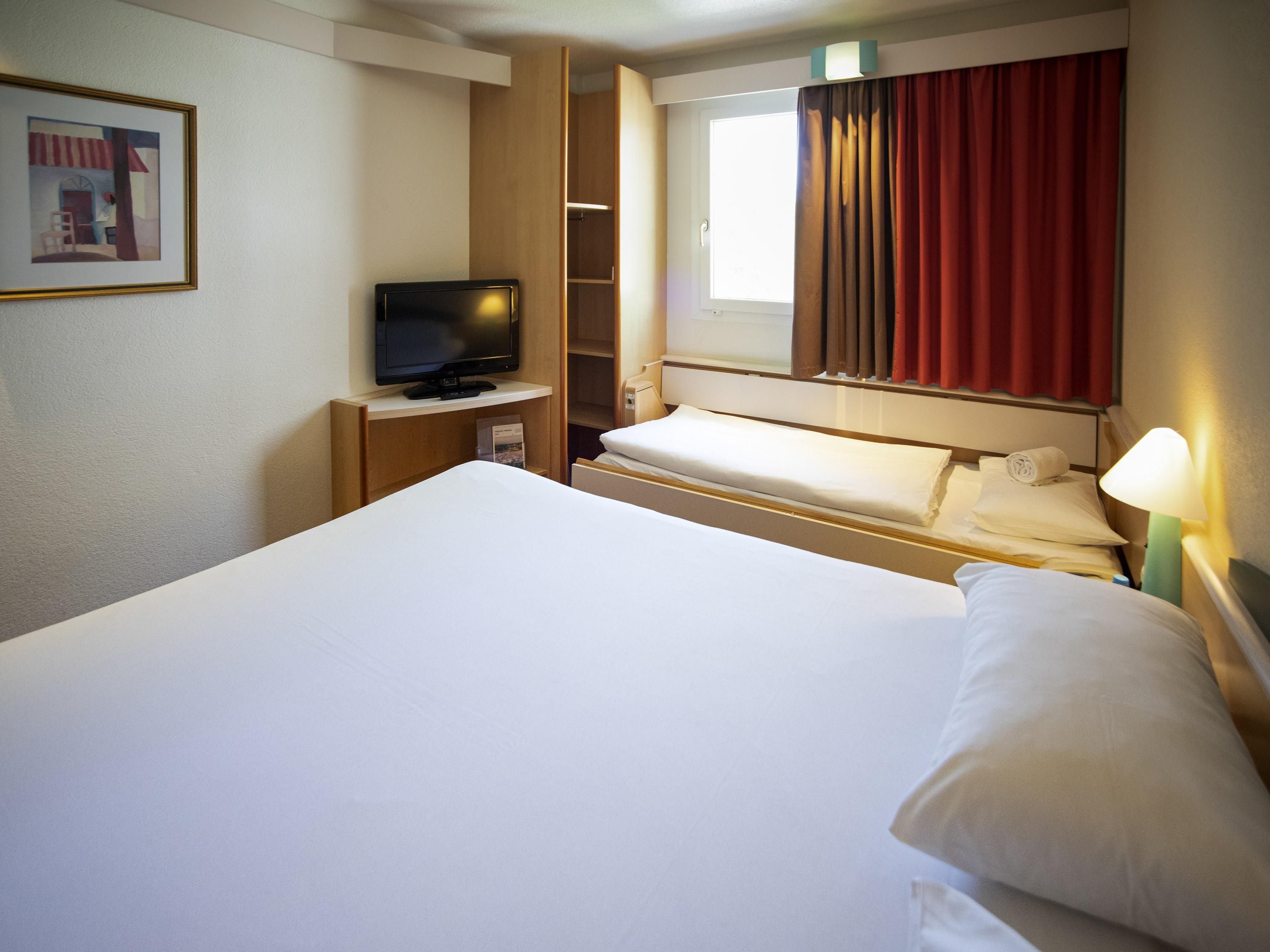 Una habitació d'hotel amb un llit gran, una televisió sobre un suport de fusta, una finestra amb cortines i un quadre a la paret.