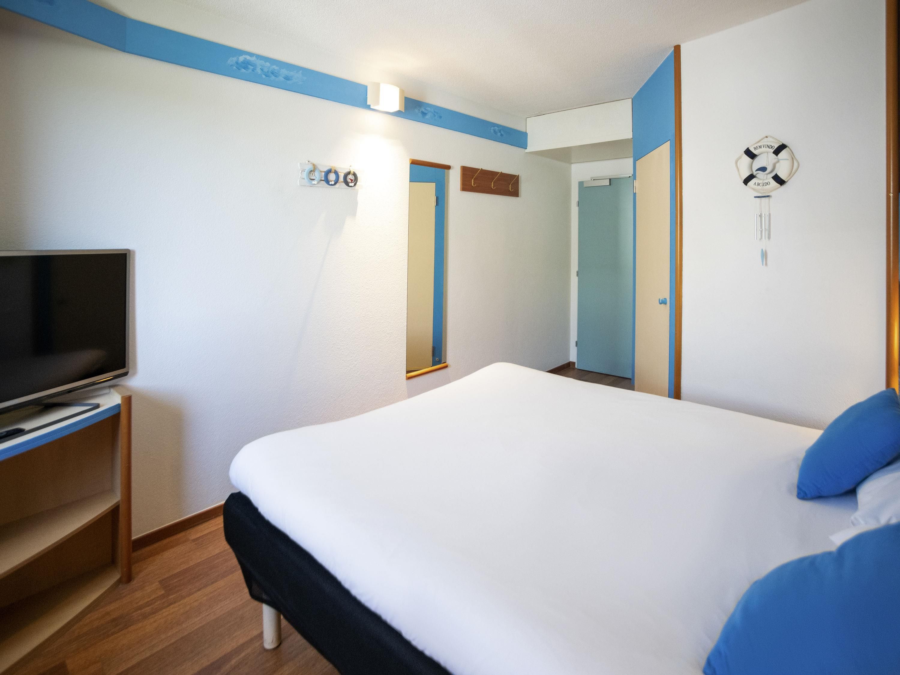 Une chambre d’hôtel avec un lit, un téléviseur posé sur un meuble, une porte et une horloge murale.