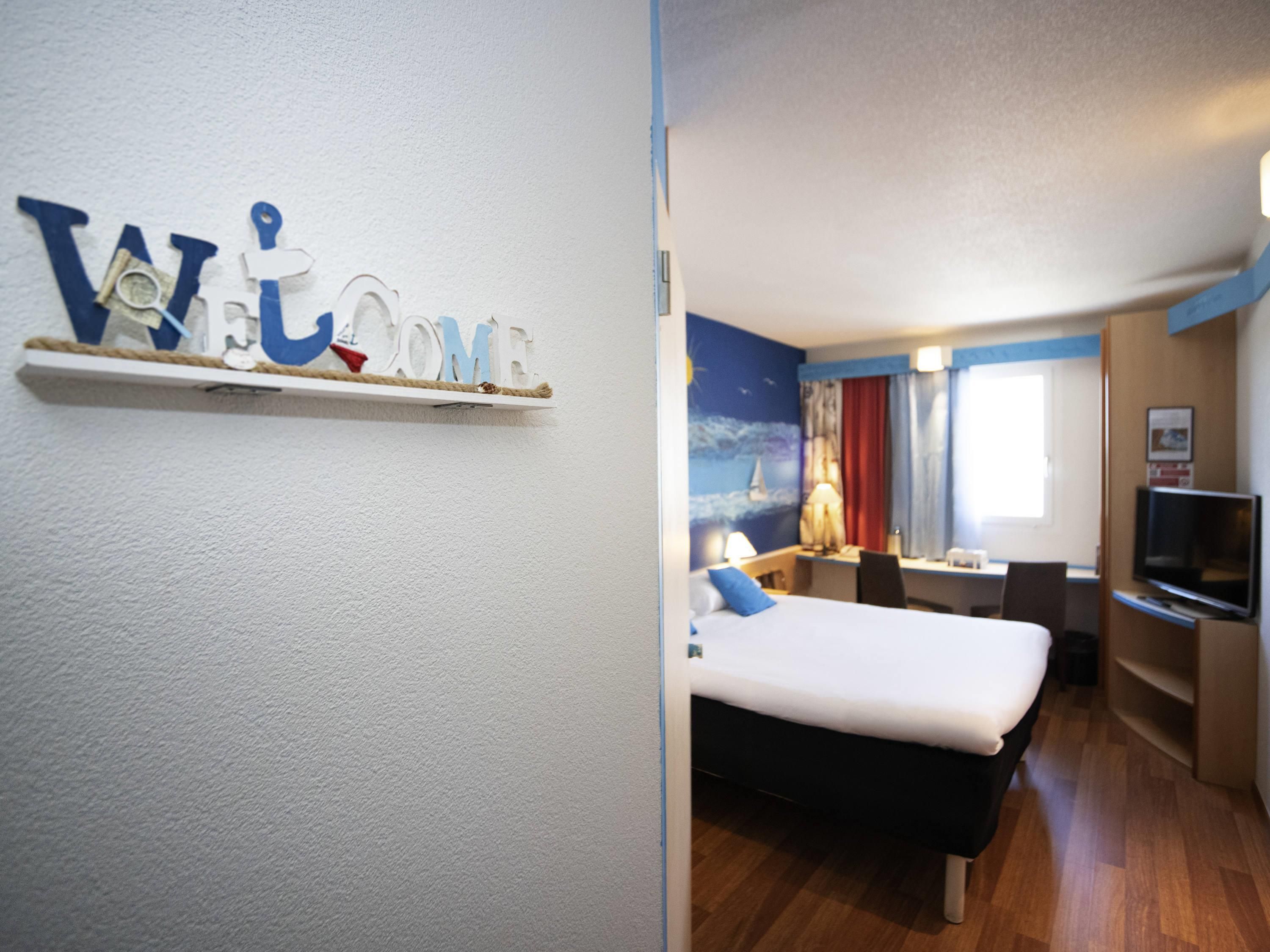 Ein Hotelzimmer mit Bett, Schreibtisch, Stuhl, Fernseher und einer an der Wand befestigten Ablage mit einer Willkommensanzeige.