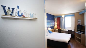 Ein Hotelzimmer mit Bett, Schreibtisch, Stuhl, Fernseher und einer an der Wand befestigten Ablage mit einer Willkommensanzeige.
