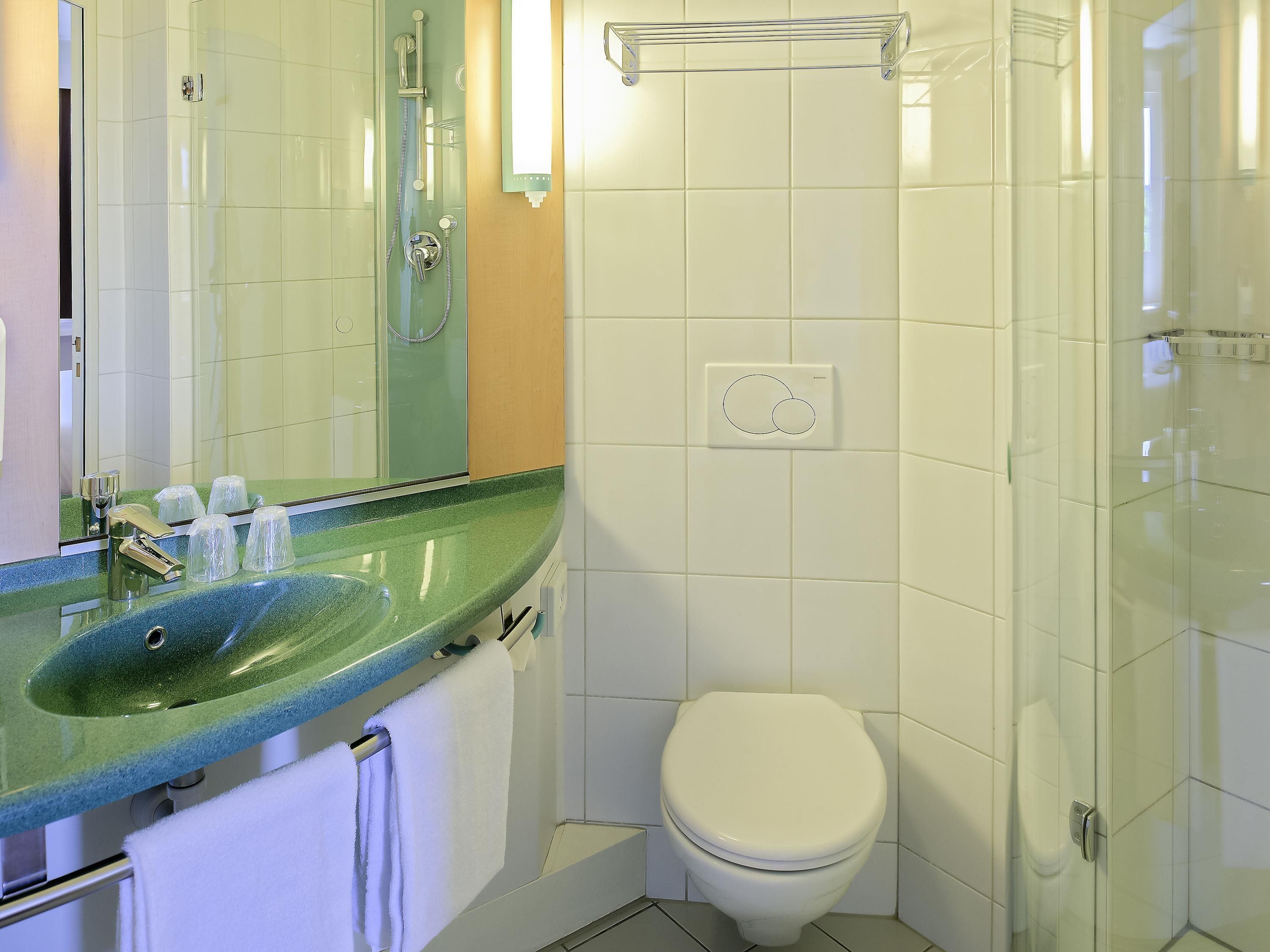 A bathroom with a glass shower enclosure, a white toilet, a sink with a green countertop, and a mirror.