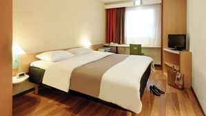 Habitación de hotel con cama, mesita de noche, lámpara, televisor, escritorio, silla y zapatero.