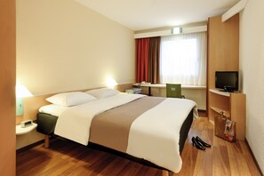 Habitación de hotel con cama, mesita de noche, lámpara, televisor, escritorio, silla y zapatero.