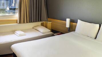 Ein Hotelzimmer mit einem großen Bett, einem kleineren Bett, einem Nachttisch, einer Lampe und einem Fenster mit Vorhängen.