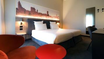 Una camera d'albergo con un letto, un tavolo rotondo rosso, una sedia, una scrivania e un'opera d'arte raffigurante un panorama urbano.