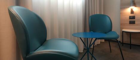 Habitación moderna con sillas color turquesa y una pequeña mesa redonda, una lámpara fijada a la pared y vistas de un edificio a través de cortinas translúcidas.