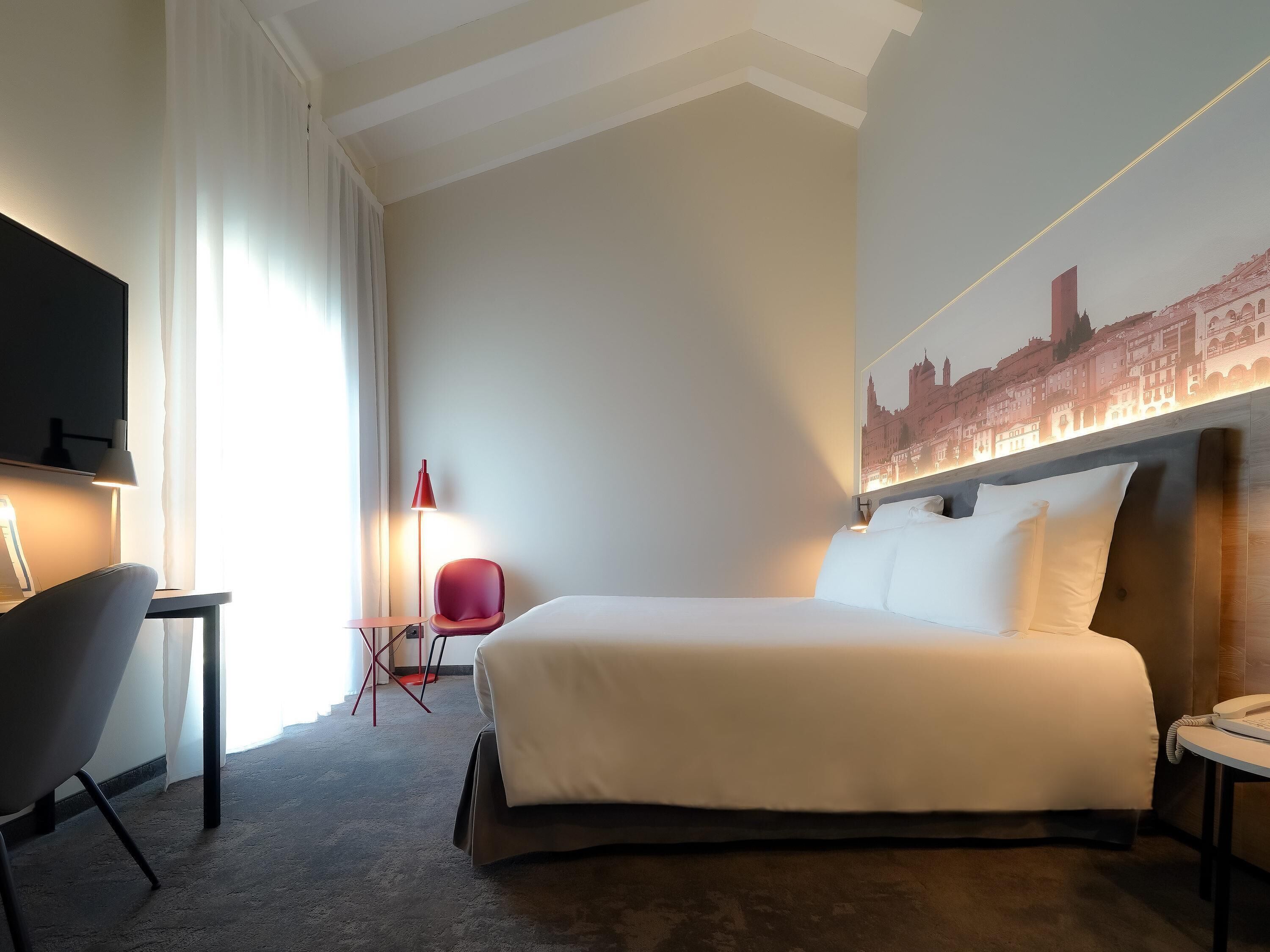 Une chambre d’hôtel équipée d’un lit, d’un bureau, d’une chaise, d’une télévision et d’un mur orné d’une fresque représentant un paysage urbain.