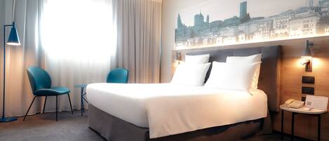 A hotel room with a large bed, two chairs, a small table, and a cityscape wall art.