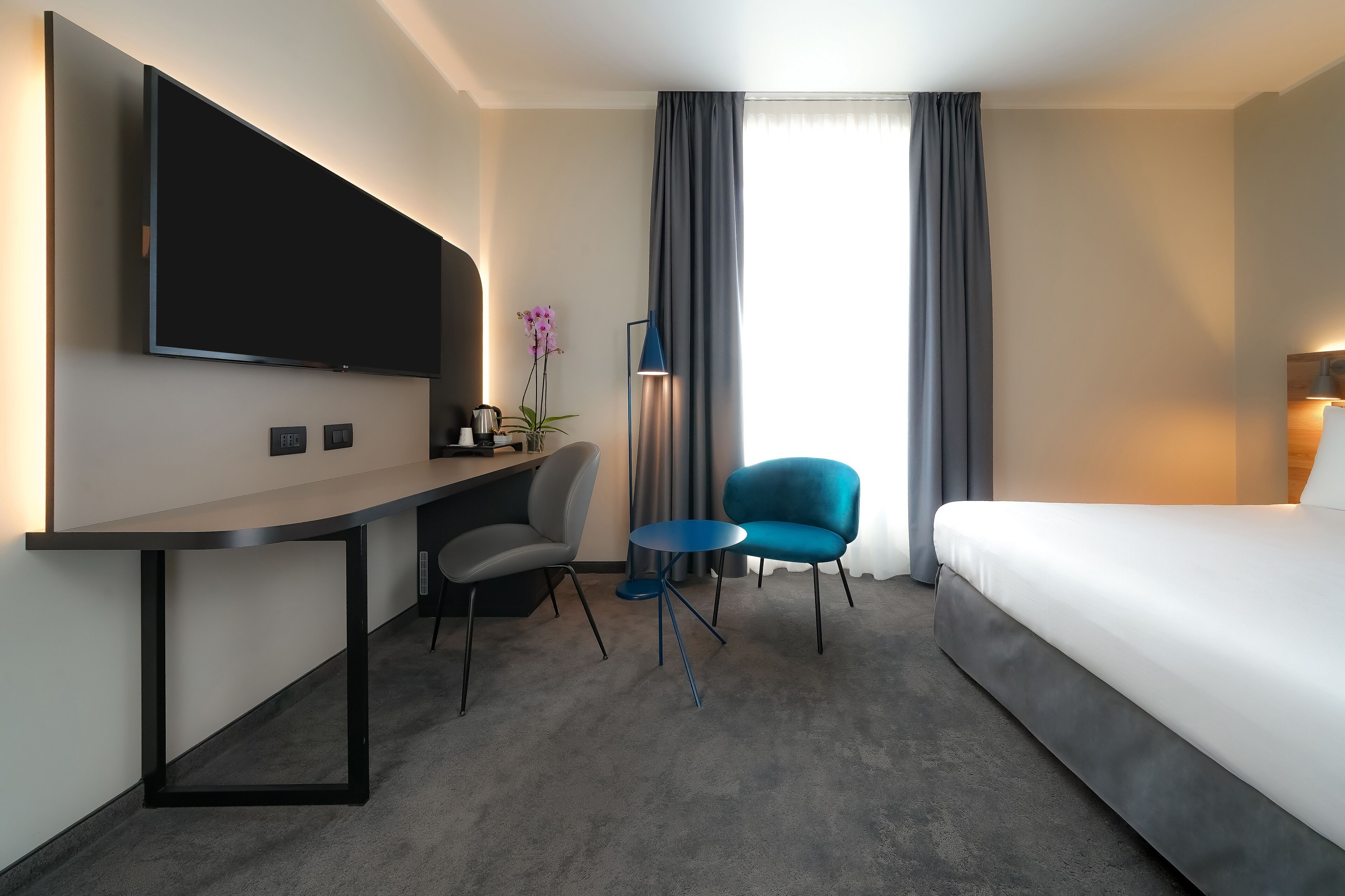 Ett hotellrum med en säng, ett skrivbord med en platt-tv, en stol, ett litet runt bord och en orkidé i en kruka.