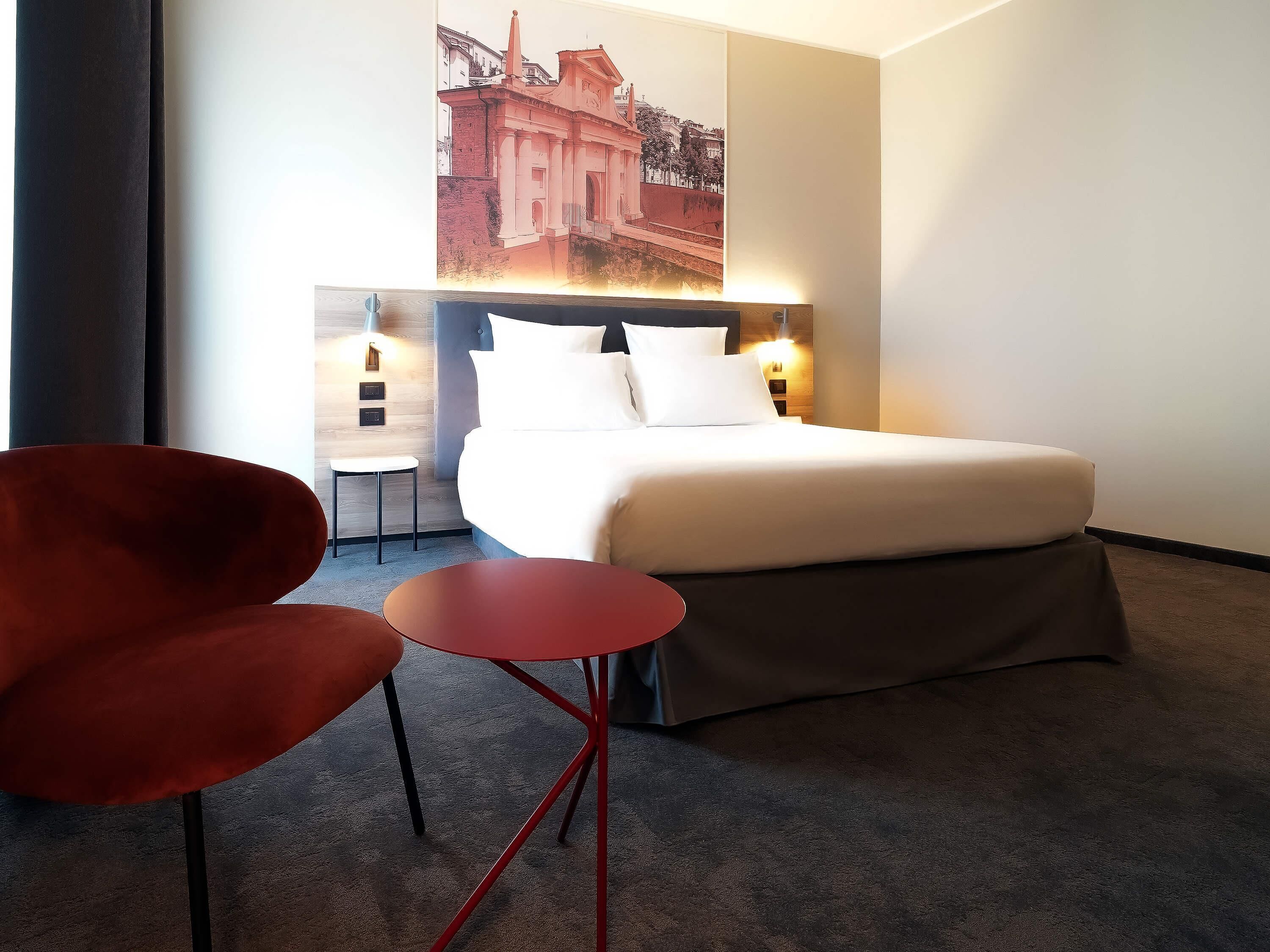 Une chambre d’hôtel avec un lit, une chaise rouge, une petite table et un lampadaire fixé au mur.