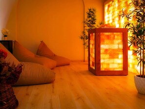 Una habitació acollidora amb terra de fusta, zona de seients amb coixins, taula auxiliar de fusta i paret amb un lluminari càlid.