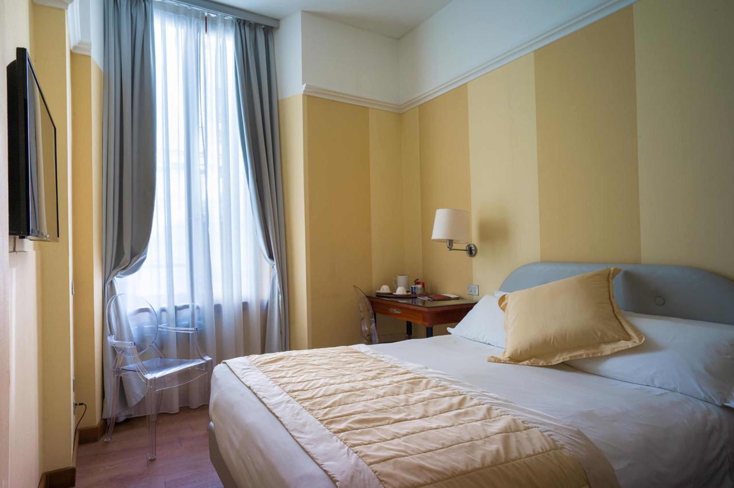 A hotel room with a bed, a desk, a chair, a TV, and a window with curtains.