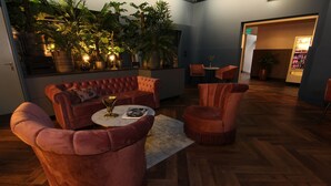 A modern lounge area with a plush red sofa, matching armchairs, and a small round table with a glass. The space is furnished with wooden flooring and features a variety of potted plants and a wall-mounted shelf with decorative items. - Savoy Hotel Amsterdam (Amsterdam)
