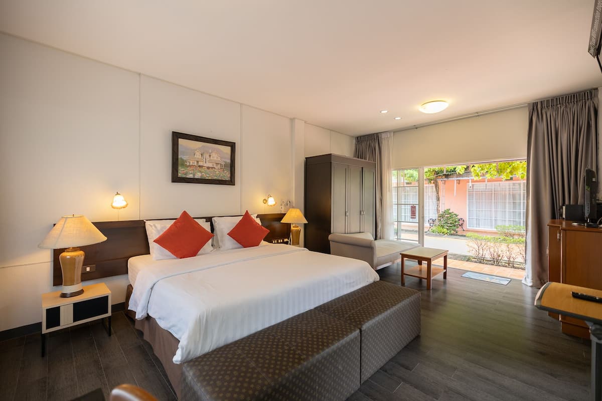 A hotel room with a large bed, two bedside lamps, a nightstand, a bench, a desk, a wardrobe, and a window with curtains.