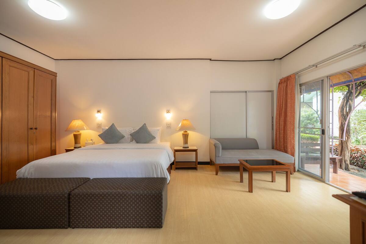 A hotel room with a bed, two bedside tables with lamps, a sofa, a small table, and a sliding glass door leading to an outdoor area.