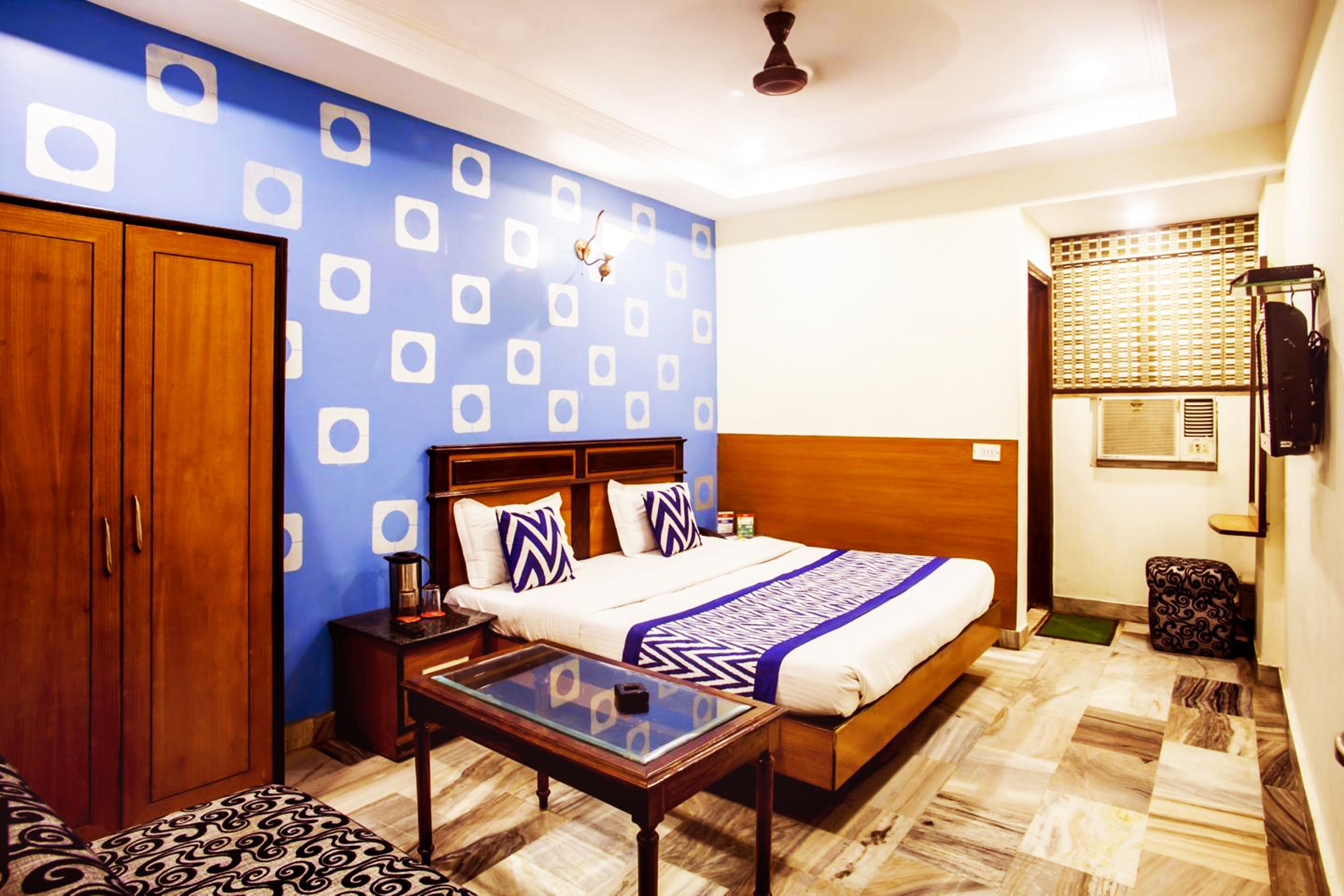A bedroom with a bed, wooden wardrobe, bedside table, and patterned ottoman.