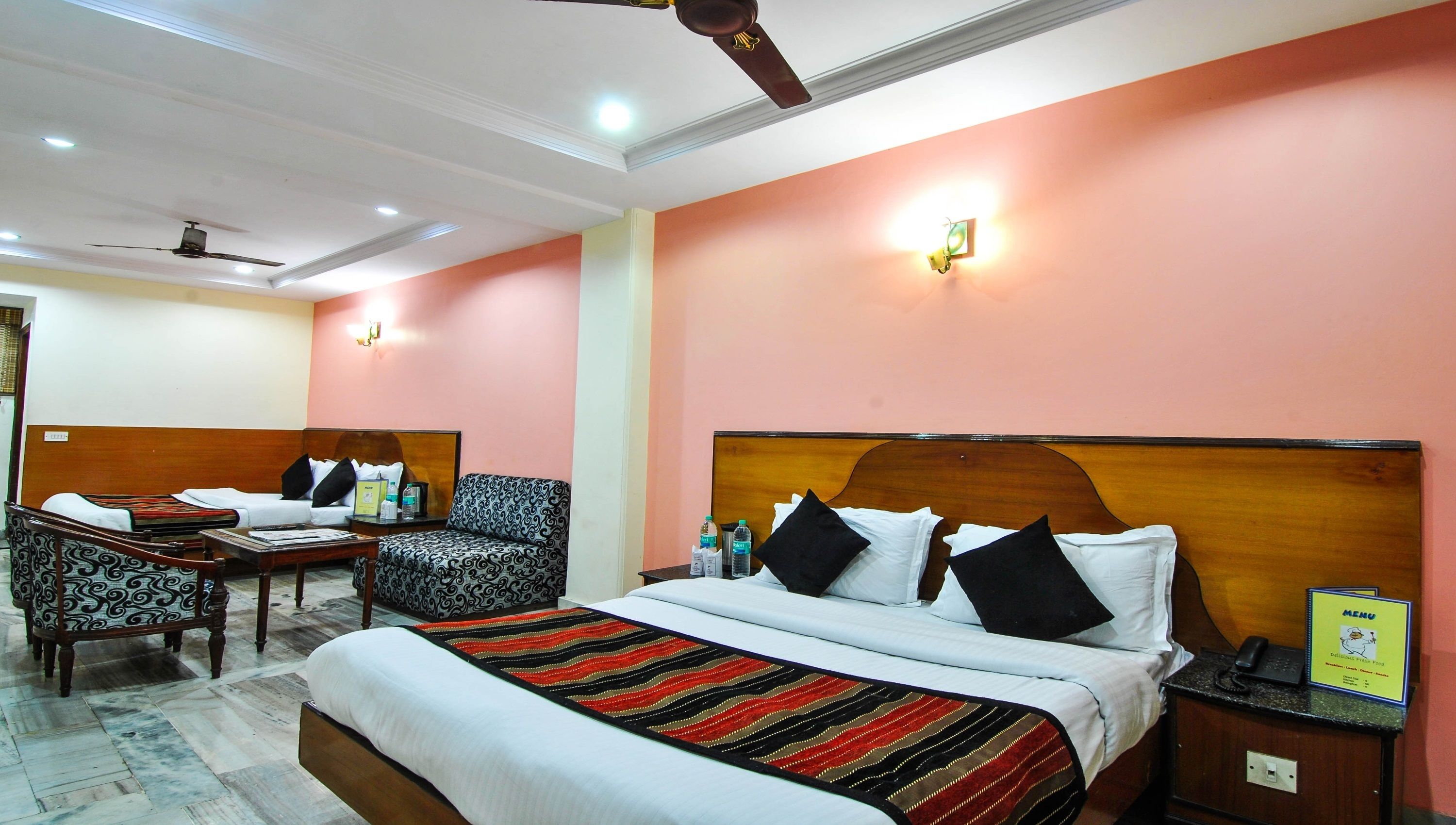 A hotel room with a large bed, a sofa, a desk, and a chair. There is a ceiling fan, a wall-mounted light, and a small table with a phone and a menu sign.