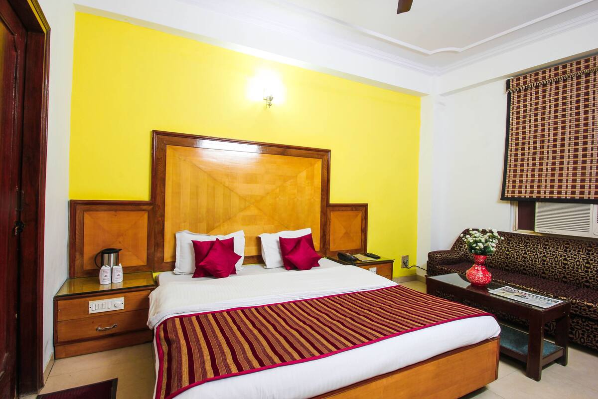 A hotel room with a large bed, a wooden headboard, a bedside table with a lamp, a sofa, and a window with blinds.