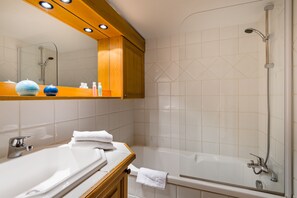 A bathroom with a white sink, mirror, and shower. There are towels on the counter and a wooden shelf above the sink.