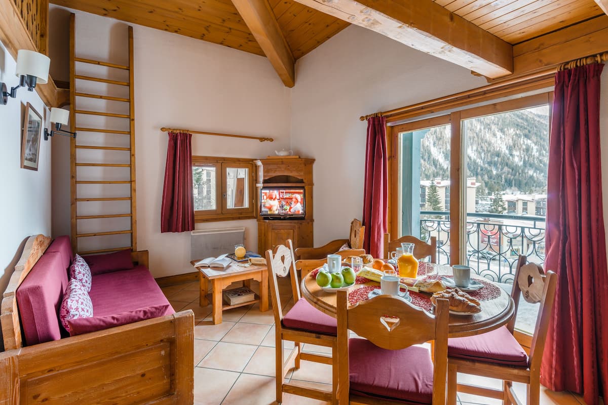 Ein gemütliches Zimmer mit Bett, einem Esstisch mit Frühstück, einem Fernseher und einem Balkon mit Bergblick.