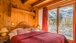 A wooden cabin bedroom with a large bed, a nightstand with a lamp, a shelf with decorative items, and a balcony with a mountain view. - Residence  Le Cristal d'Argentière (Chamonix-Mont-Blanc)