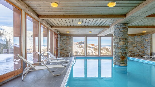 An indoor swimming pool with stone walls and wooden beams, surrounded by lounge chairs and large windows offering a view of a snowy landscape. - Résidence Lagrange Vacances Aspen (La Plagne-Tarentaise)