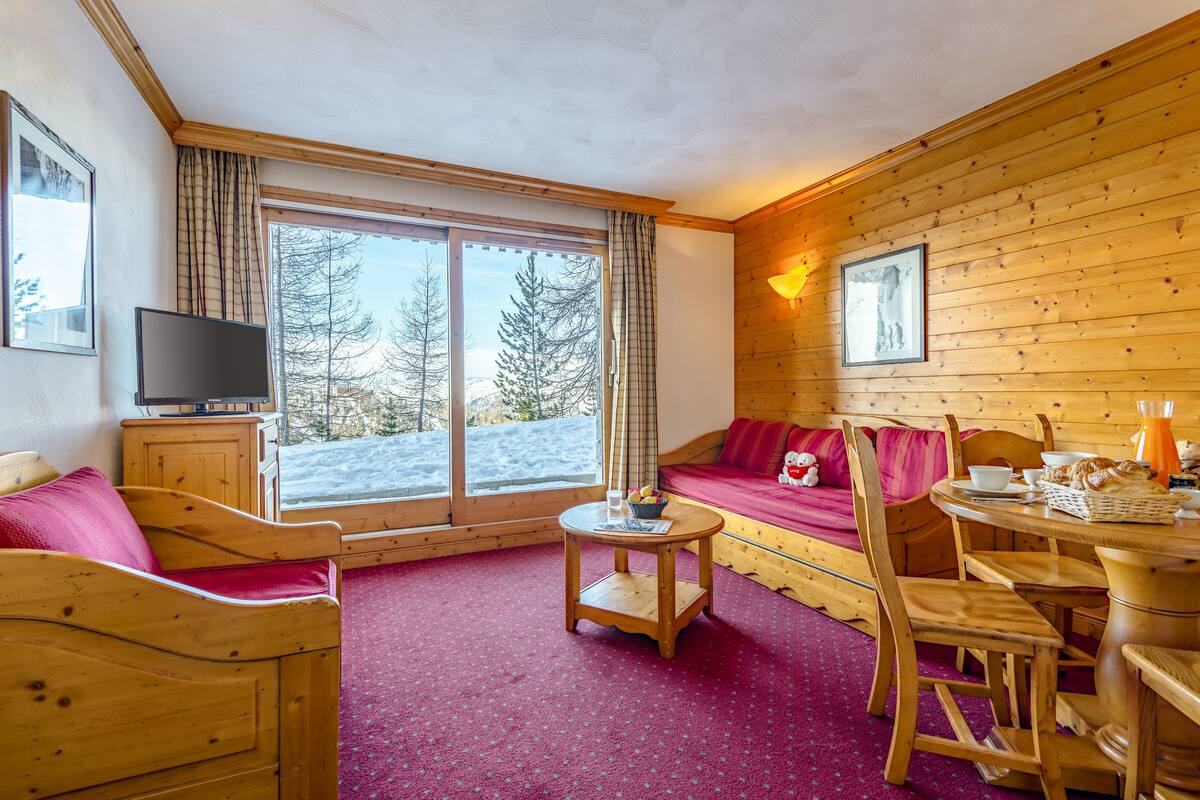Une pièce chaleureuse avec un décor intérieur en bois, une grande fenêtre donnant sur les arbres, un canapé, une table avec un saladier de fruits et un téléviseur posé sur un meuble.