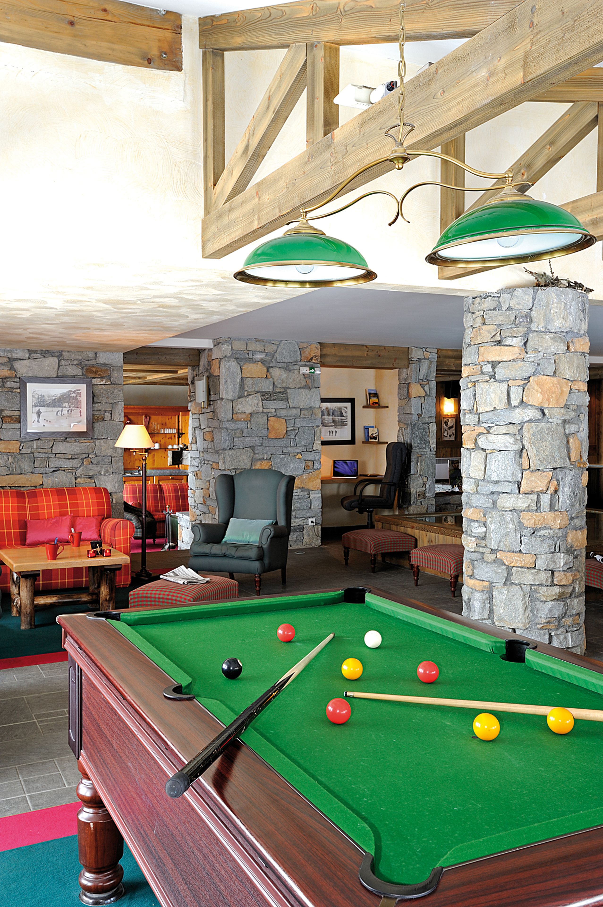 A pool table with balls, a green sofa, a chair, and a lamp in a room with stone walls and wooden beams.