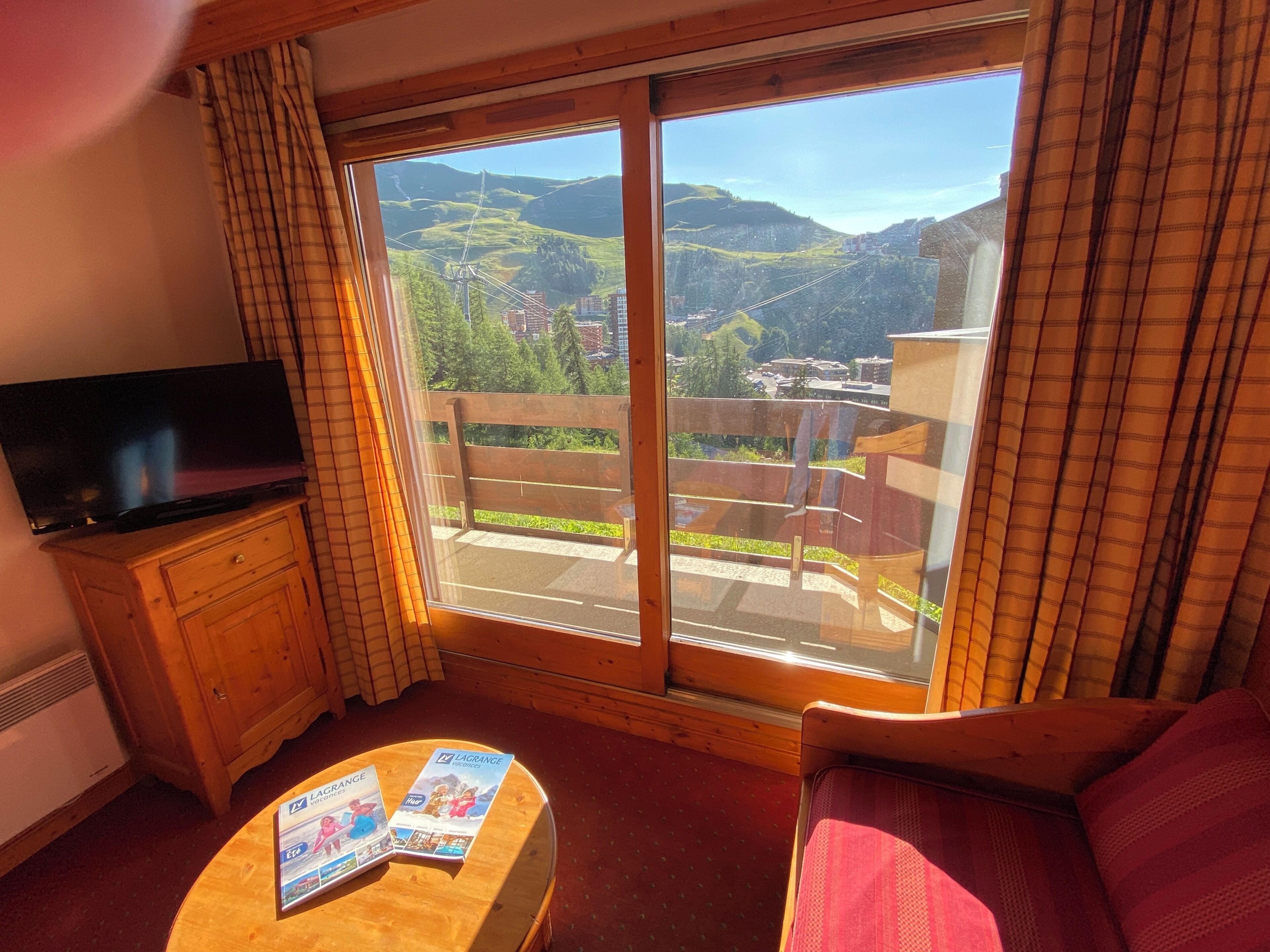 A room with a television on a wooden cabinet, a red sofa, a wooden coffee table with brochures, and a large sliding glass door leading to a balcony with a view of green hills and buildings.