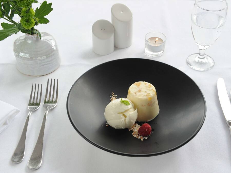 A dessert plate with a scoop of ice cream and a cube of dessert on a black plate, accompanied by a fork, knife, and a glass of water.