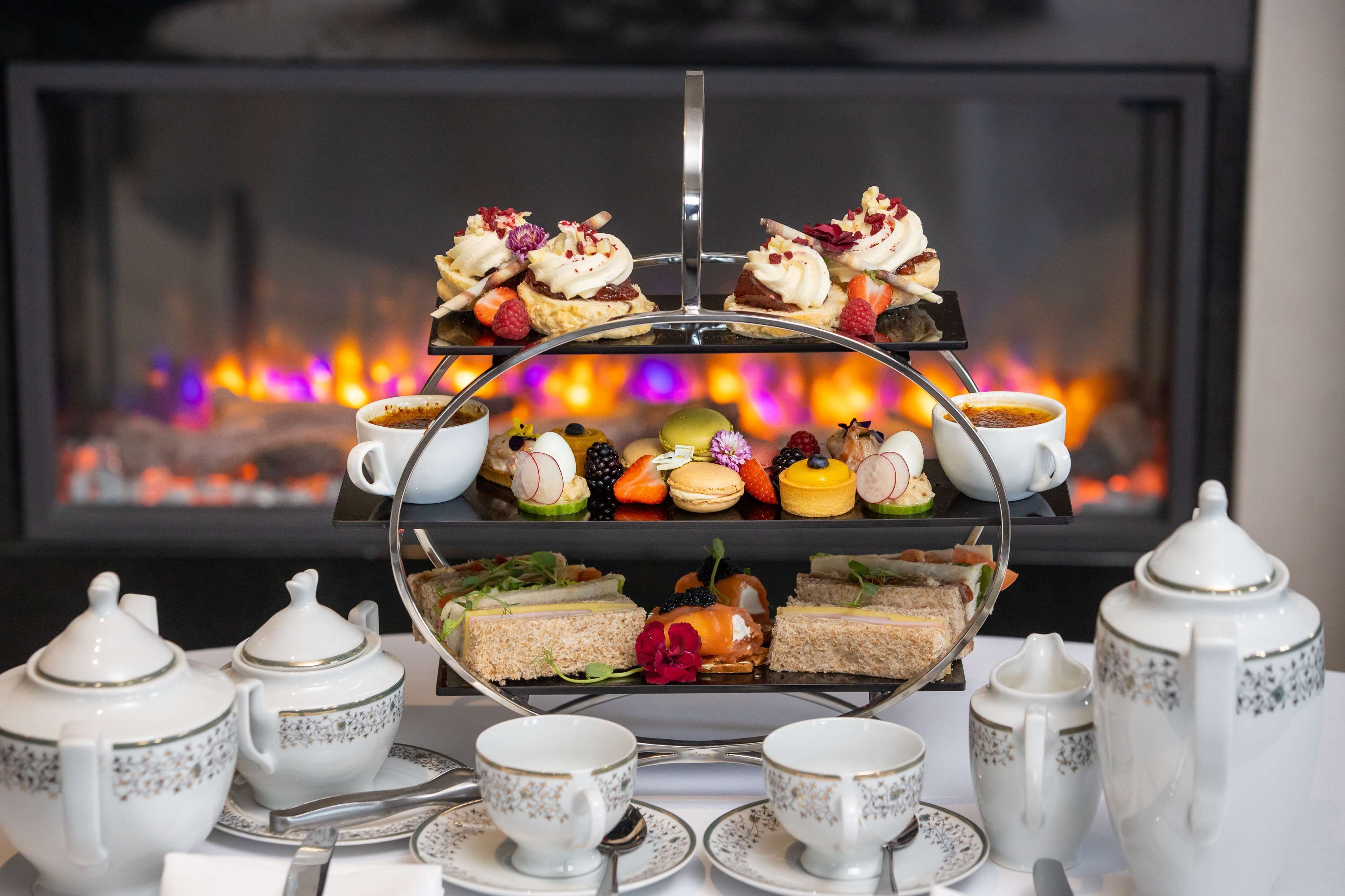 Ein mehrstufiges Desserttablett mit verschiedenen Gebäcken und Früchten, begleitet von Teesets und Teekannen, vor einer Kaminfeuerstätte.