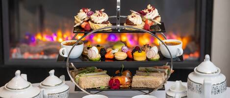 A three-tiered dessert tray with various pastries and fruits, accompanied by tea sets and teapots, set against a backdrop of a fireplace.