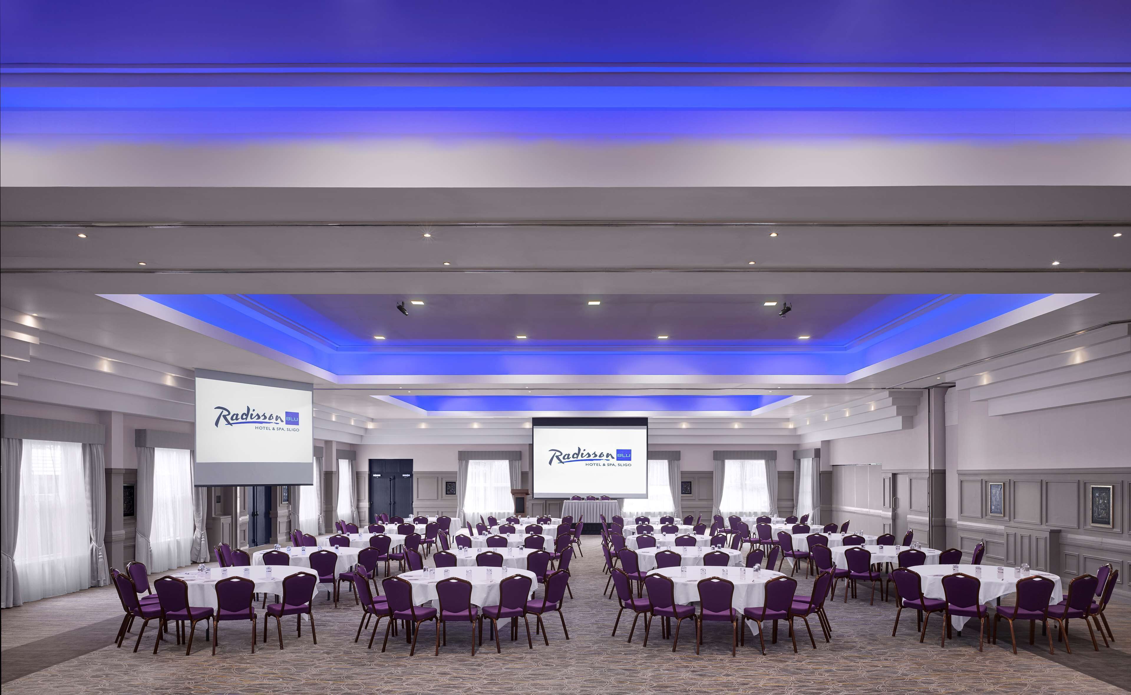 Un salón de conferencias con varias mesas y sillas, pantallas grandes mostrando el logotipo de Radisson, y un diseño moderno de techo.