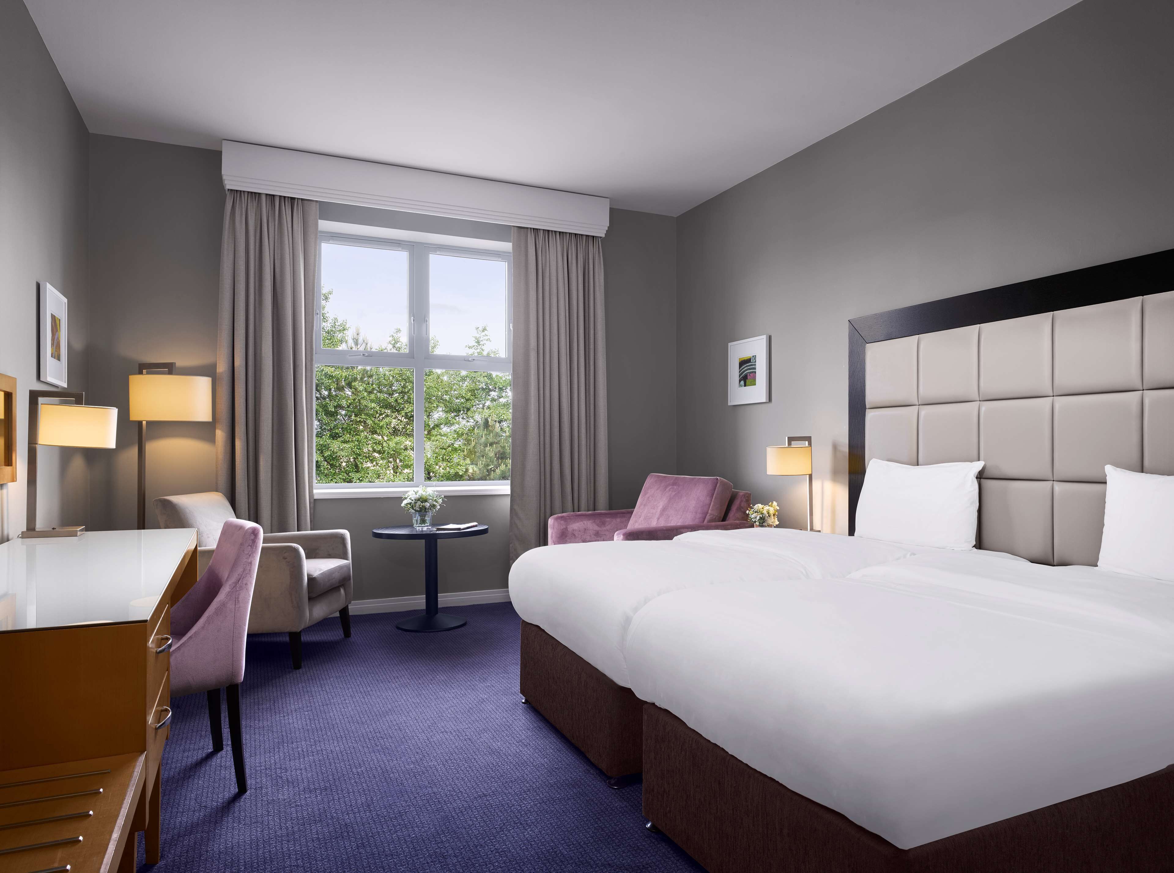 Une chambre d’hôtel avec deux lits, un bureau, une chaise, une petite table et une fenêtre avec des rideaux.